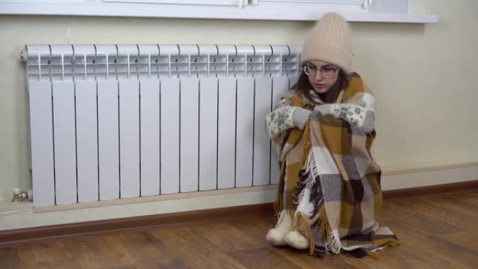 一个年轻的女人坐在暖气片旁边的地板上，身上盖着一条毯子。女孩还没有付暖气费，坐在家里穿着暖和的衣服。