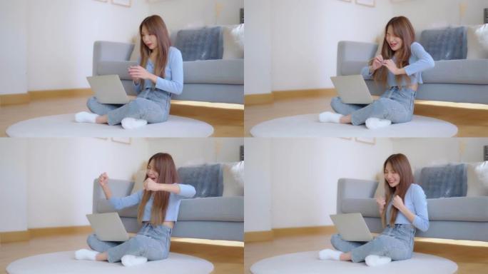 年轻的亚洲女性精神振奋，坐在靠近沙发的地板上，在家用笔记本电脑工作。兴奋的女性获胜者在浏览笔记本时庆