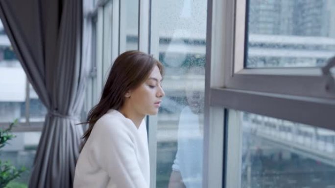 年轻的亚洲妇女独自在家思考孤独或悲伤坐在窗边，带着孤独的表情看着外面