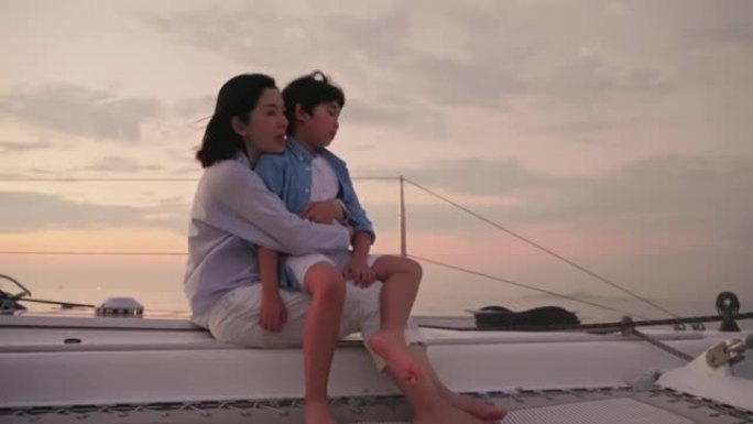 快乐的母亲拥抱儿子在白色游艇上观看假日日落