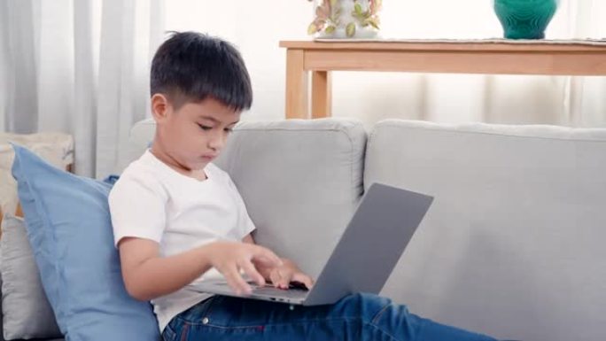 4K，亚洲男孩坐在打开的笔记本上，早上在线学习，坐在客厅沙发上，看起来决心学习，下课了，他们坐下来做