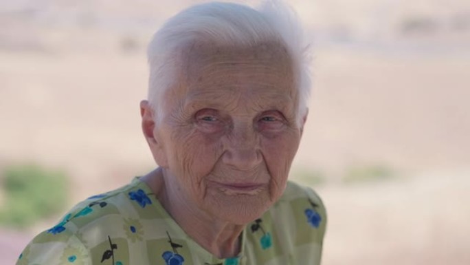 满脸皱纹的白人老妇用灰色的眼睛看着相机的头像。灰发退休人员在阳光明媚的夏日度假时在户外摆姿势。旅游和