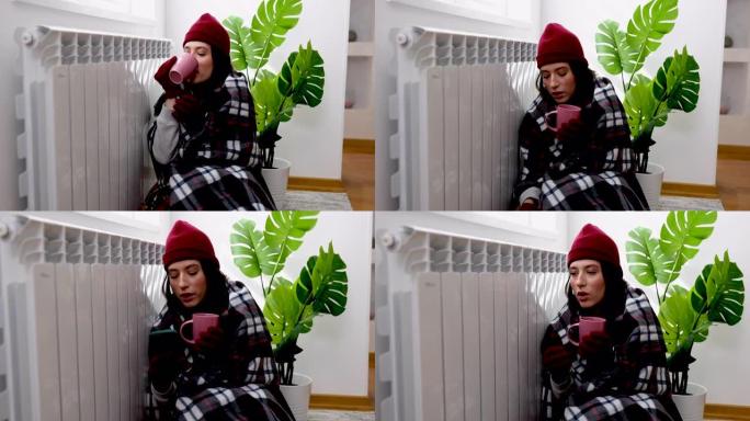 一个穿着暖和衣服的女人在暖气片和使用电话的同时，在家里发抖，戴着毯子，冻着。