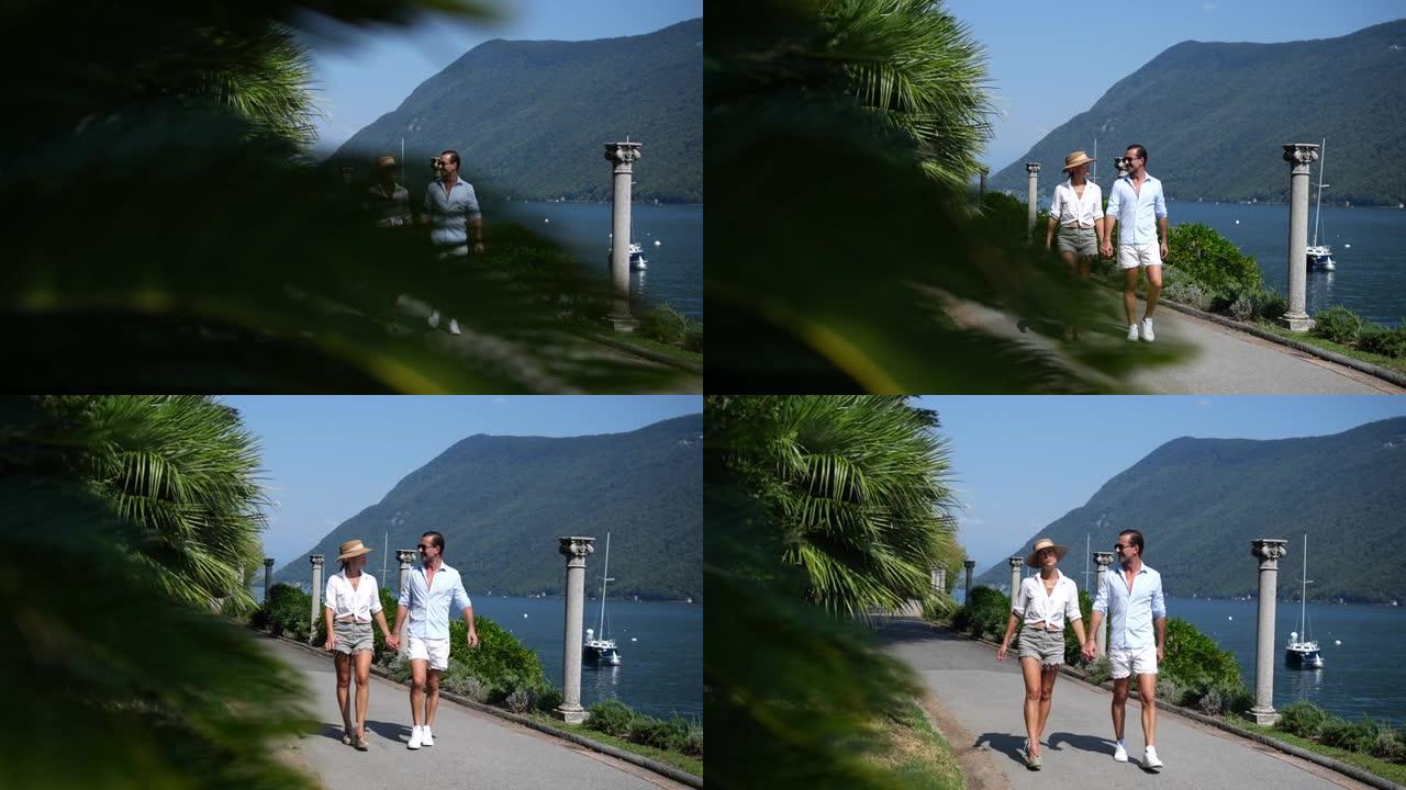 男人和女人手牵着手在湖边穿过公园，穿着休闲的夏季服装，可以看到后面的山脉