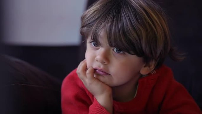 一个小男孩特写脸，用手握下巴在镜头下观看动画片。小孩的肖像盯着躺在沙发上的屏幕媒体