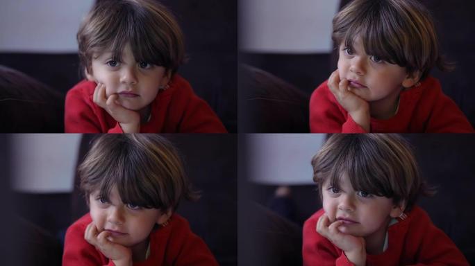 一个小男孩特写脸，用手握下巴在镜头下观看动画片。小孩的肖像盯着躺在沙发上的屏幕媒体