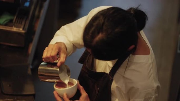4k亚洲男子咖啡师在浓缩咖啡中倒入泡沫牛奶，在咖啡杯中煮咖啡拿铁。