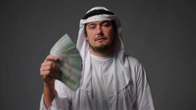 穿着传统衣服的成功的阿拉伯酋长在灰色背景上手里拿着钱。富有的阿拉伯人概念
