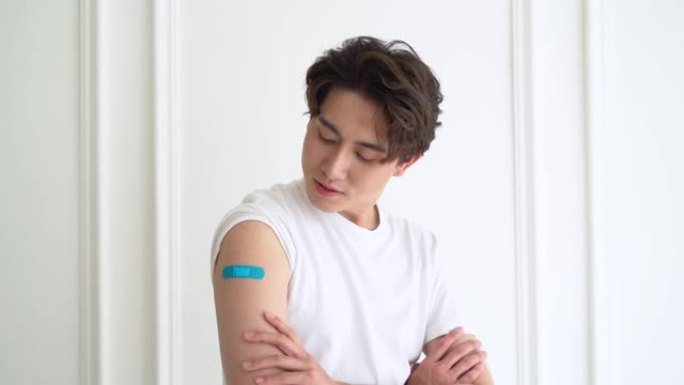 年轻的亚洲男子患者的肖像显示肩膀上的膏药表明注射后，用新型冠状病毒肺炎病毒接种疫苗后感到满意，并看着