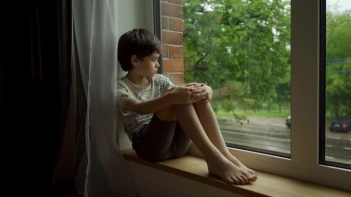 下雨时，一个男孩坐在潮湿的窗户上，看着街道