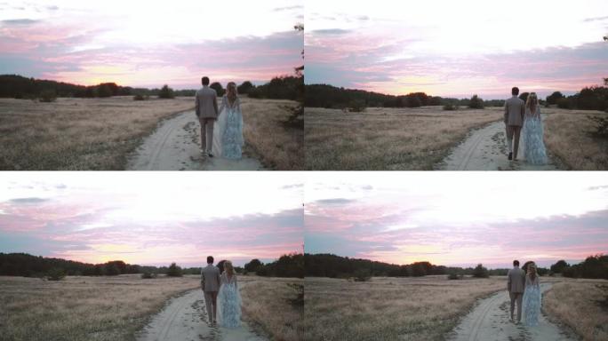 新娘和新郎的后视图在日落时穿过干草场，天空中有粉红色的云彩。穿着波西米亚风格连衣裙的新娘。日落时相爱