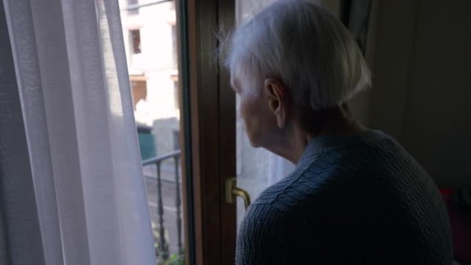 沮丧的孤独的高加索老妇人站在阳台的门口，看着夏天的街道。悲伤的高级女性退休人员想独自在家度过一天。