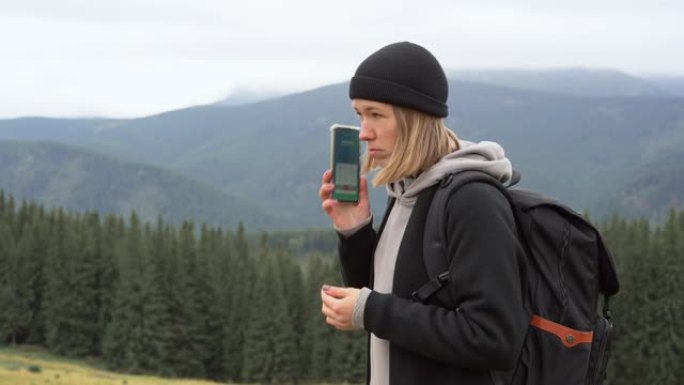 在山顶上，一位戴着帽子，背着背包的年轻金发女子拿出手机，拨打电话。移动网络覆盖概念在世界任何地方。