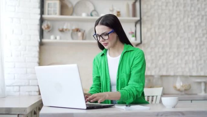 戴眼镜的年轻女子坐在轻便的厨房里，对笔记本电脑有问题感到恼火。心烦意乱的女孩不能工作，因为系统错误，