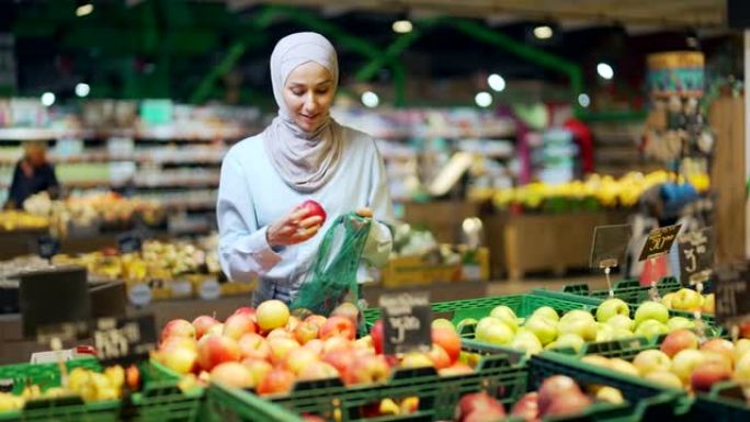 戴着头巾的年轻穆斯林妇女在杂货店超市或市场的架子上选择和采摘水果，然后将它们放在生态袋中，女顾客扔苹