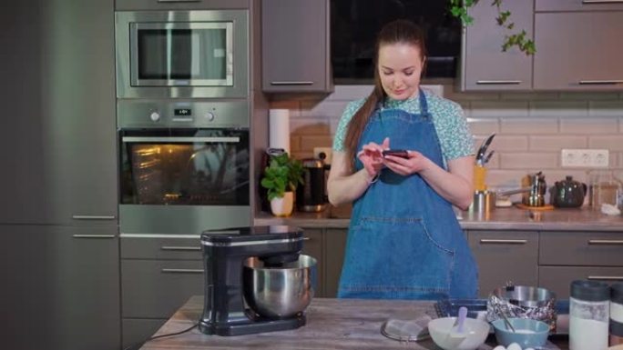 一个年轻漂亮的女人站在厨房里，微笑着看着智能手机里的东西。在她面前是一个工作的烹饪搅拌机，并布置了烘