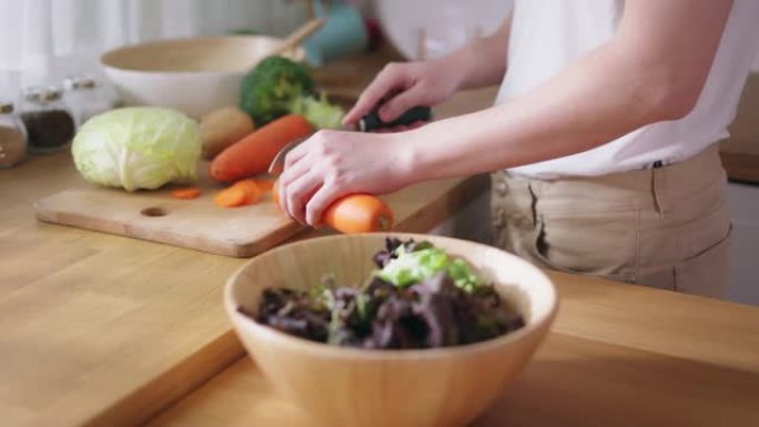 穿着白色t恤的年轻人的特写镜头在家里的厨房里切白菜和胡萝卜等生蔬菜。素食主义者做沙拉。