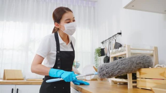 亚洲清洁服务女工在家厨房打扫卫生。漂亮的女管家清洁工戴口罩，用鸡毛掸子擦拭凌乱的柜台做家务或家务。