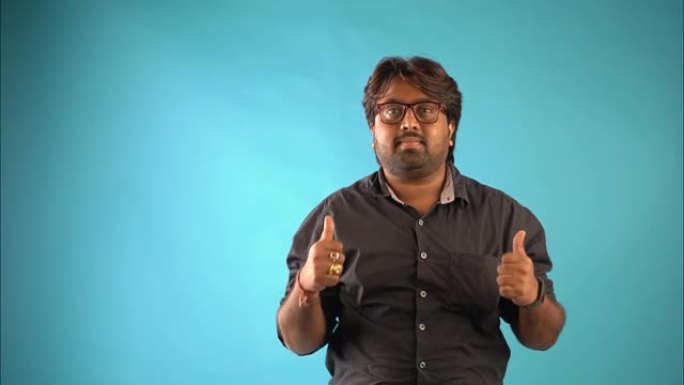一名穿着黑色衬衫的印度男子站在孤立的蓝色背景中竖起大拇指