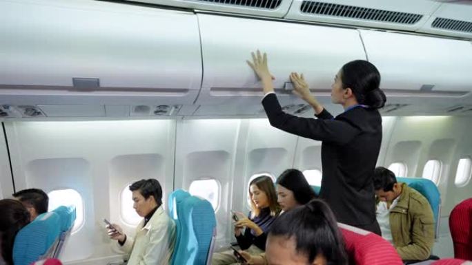 穿着制服的美丽的亚洲空姐帮助乘客将背包和行李放在头顶的车厢中并关闭，而机舱中的乘客则坐着，使用智能手