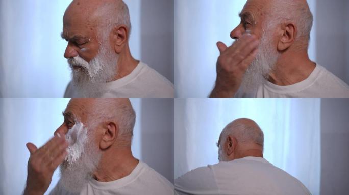 侧视聚焦的白人老人在慢动作的脸颊上涂抹白色剃须泡沫。集中的老退休人员在室内浴室设计胡须。