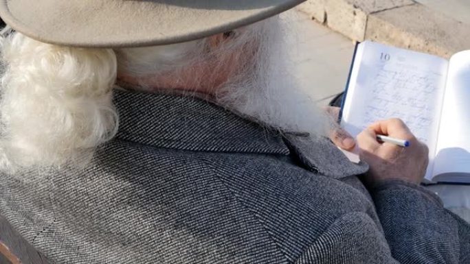 老人身后的俯视图，留着灰色胡须和白色辫子，头上戴着帽子，灰色外套坐在户外的长凳上，用笔在笔记本上写笔