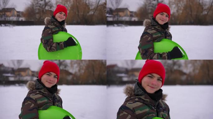放大微笑的十几岁男孩在户外用绿色雪橇慢动作摆姿势的肖像。快乐放松的高加索英俊少年看着相机站在寒冷的冬
