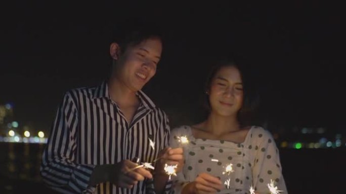 这对幸福的夫妇手持烟火灯，晚上在海滩上享受烟花汇演。