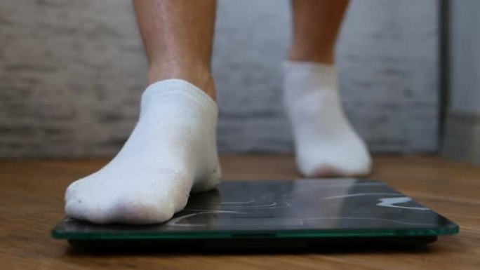 穿着白色袜子的男人的脚站在地板上。一个人在秤上称重以控制自己的体重。减肥概念。称量体重秤上的男性身体