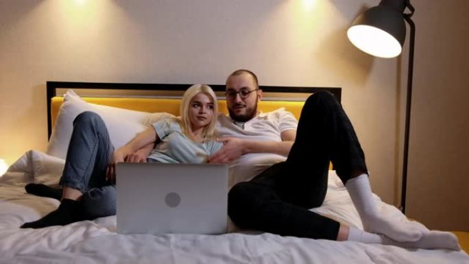 快乐轻松的年轻夫妇躺在沙发上，在笔记本电脑上观看电影，照片或电视连续剧
