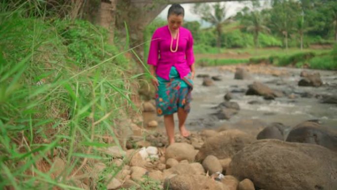 亚洲女人穿着粉红色的连衣裙和绿色的裙子在河边散步，化妆时石头很多