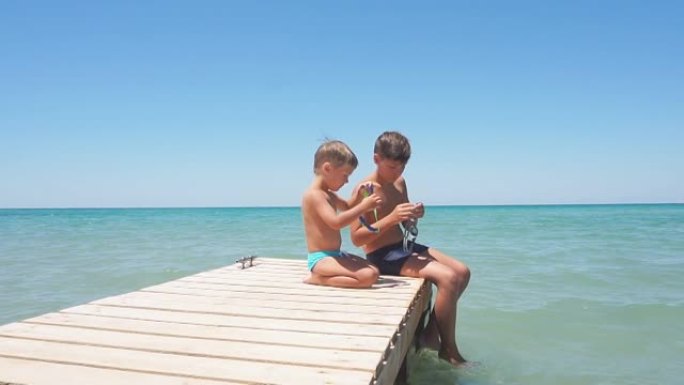 在阳光明媚的炎热日子里，孩子们坐在蔚蓝的大海背景下的劣质码头上，男孩想在海里游泳，所以戴水镜