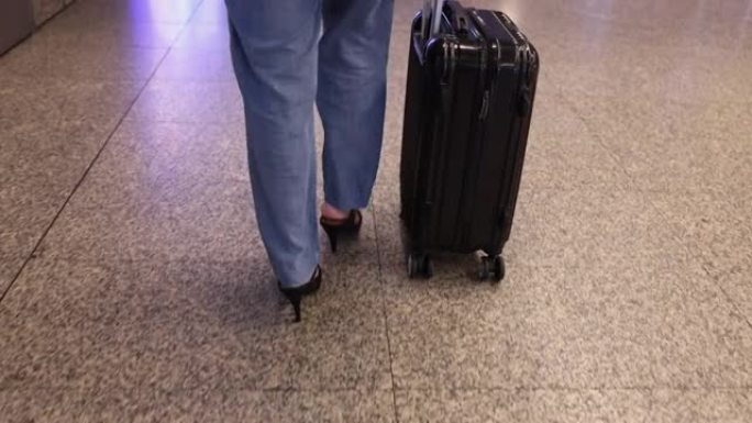 女人穿着牛仔裤长裤，黑色高跟鞋，黑色手提箱沿着机场航站楼行走，腿的底部视图。