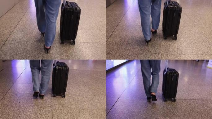 女人穿着牛仔裤长裤，黑色高跟鞋，黑色手提箱沿着机场航站楼行走，腿的底部视图。