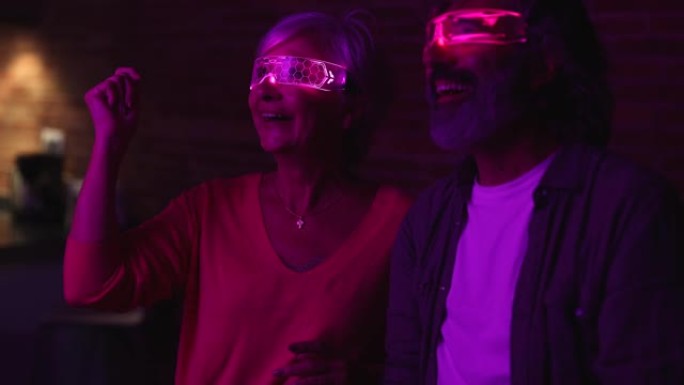 在绿光的房间里，穿着未来派霓虹灯智能眼镜的高级夫妇感到惊讶。关注白发成熟女人