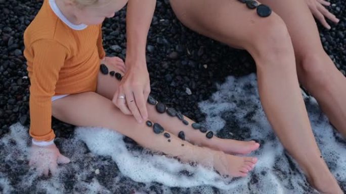 海边有黑色光滑鹅卵石。一个女人和一个小女孩坐在岸上。不时有波浪在他们身上翻滚，洗脚。妈妈把鹅卵石放在