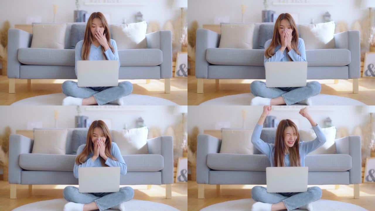 年轻的亚洲女性精神振奋，坐在靠近沙发的地板上，在家用笔记本电脑工作。兴奋的女性获胜者在浏览笔记本时庆