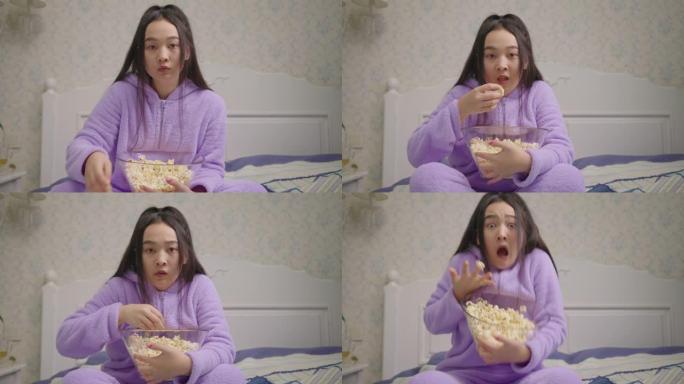 漂亮的20多岁亚洲女人看恐怖电影，独自坐在床上吃爆米花。拿着爆米花的千禧一代女人看着相机，感到害怕。