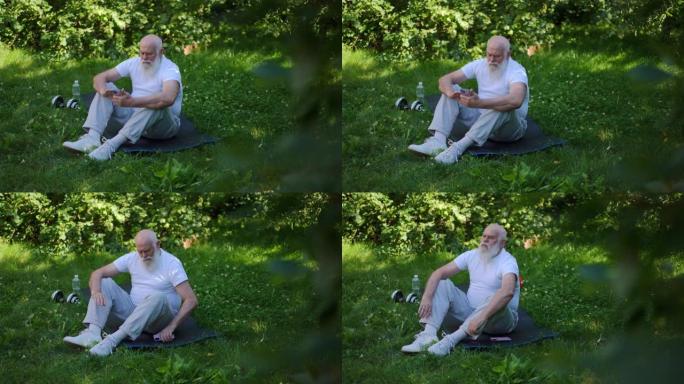 宽镜头体贴的高级运动员坐在夏季公园的运动垫上，在智能手机应用程序中滚动社交媒体。有动力的高加索老人在