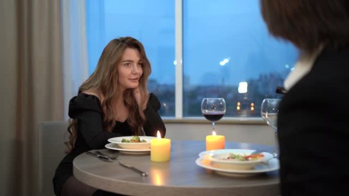 在家烛光下的浪漫晚餐，一个穿着黑色连衣裙的美女坐在桌子旁，与一个男人聊天。这家伙拿了一瓶红酒，倒进玻