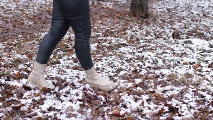 冬天森林里的女人。穿着紧身皮裤和靴子的人的腿沿着雪公园行走，相机向右移动。女人悠闲地在树上的白色湿雪