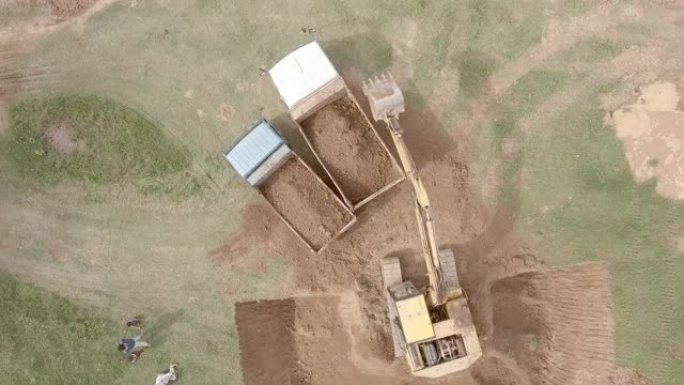 在空地上，一辆挖掘机在两辆卡车内装载泥土的高空无人机拍摄