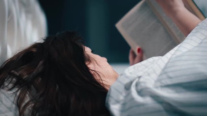 漂亮的黑发女人盖着毯子，躺在床上，晚上看书，拍摄电影