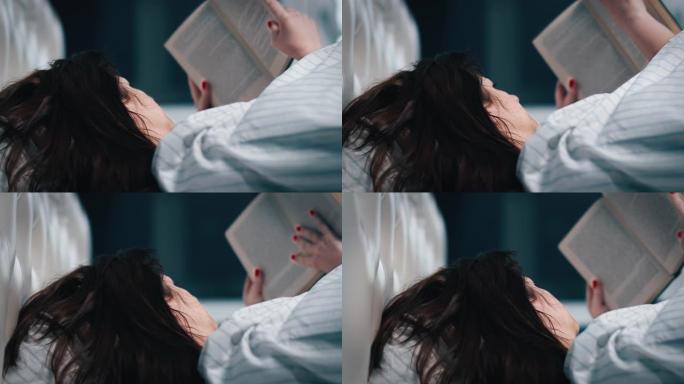 漂亮的黑发女人盖着毯子，躺在床上，晚上看书，拍摄电影