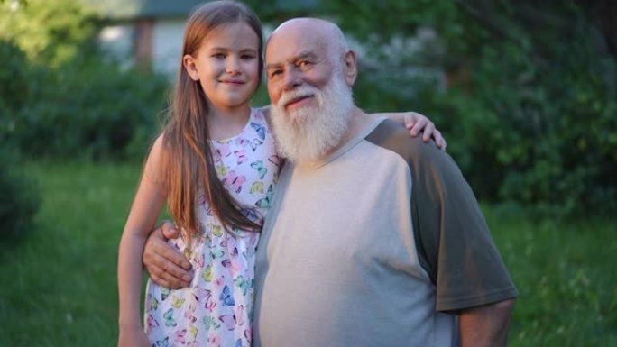 放大拥抱快乐的高加索高级祖父和孙女微笑着看着相机的肖像。积极自信的老胡子男人和迷人可爱的女孩在公园慢