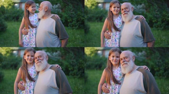 放大拥抱快乐的高加索高级祖父和孙女微笑着看着相机的肖像。积极自信的老胡子男人和迷人可爱的女孩在公园慢