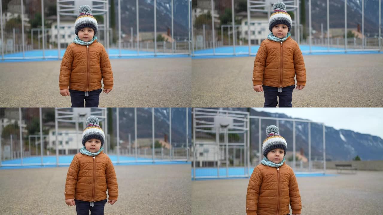 冬季，小男孩穿着豆豆围巾和夹克站在公园外面。沉思的蹒跚学步的孩子穿着暖和的衣服站在户外