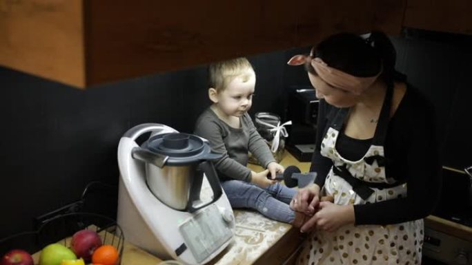 黑色厨房捏合机附近的桌子上坐着短发的小男孩婴儿蹒跚学步的俯视图，与年轻的母亲一起用黑色硅胶刮铲在面粉