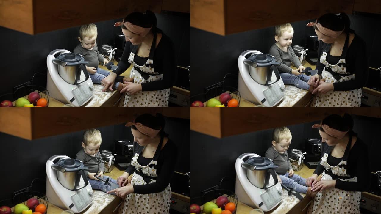 黑色厨房捏合机附近的桌子上坐着短发的小男孩婴儿蹒跚学步的俯视图，与年轻的母亲一起用黑色硅胶刮铲在面粉