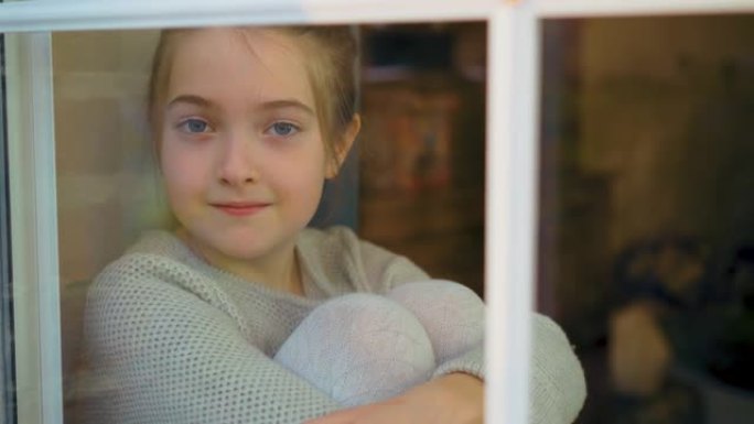 肖像可爱的小女孩微笑着从这里的家看着窗外，坐在窗台上